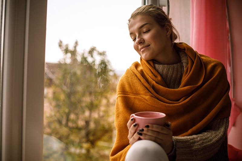 Woman holding a hot beverage in a mug, sitting by a window.